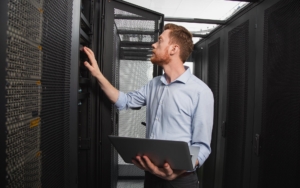 Side view of an IT worker holding laptop and looking at servers in data center