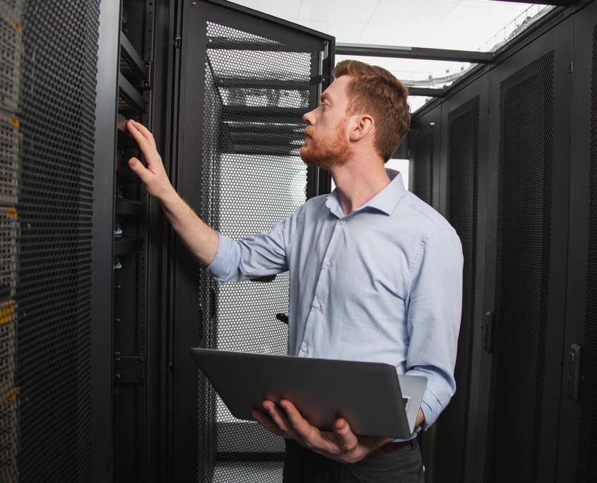 Side view of an IT worker holding laptop and looking at servers in data center