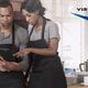 man in apron looks at tablet while woman in apron points at screen in commercial kitchen