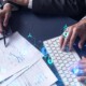 man with laptop reviews paper notes while coworker types on keyboard